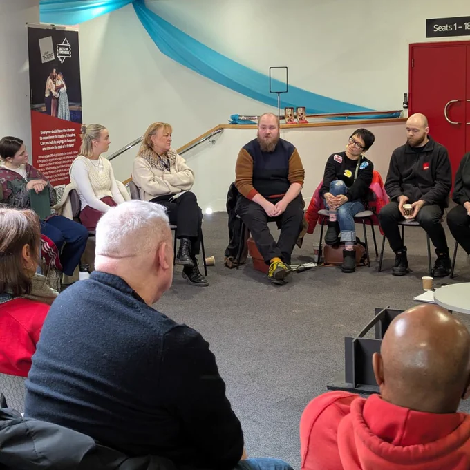 A discussion setup with people seated in a semi-circle in a room decorated with blue fabric draped across the ceiling. A man in a brown sweater speaks while others listen, with informational posters visible on the wall.