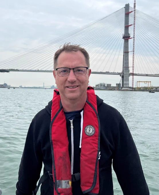Trevor is wearing glasses, a black hoodie,  and red lifejacket. He is posing in front of a body of water with a bridge in the background.