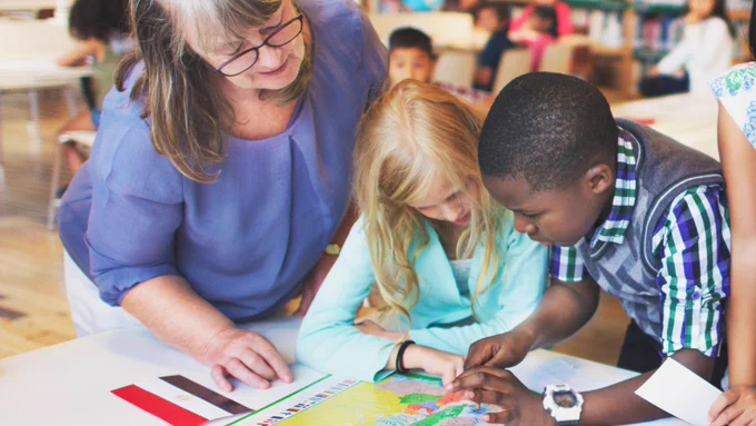 A teacher in a blue shit helping two young students look over a map on a table.