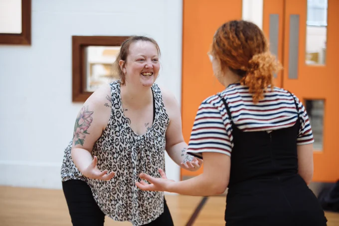 Two people stand opposite each other in a brightly lit room. They grin at each other, in the midst of what appears to be a game of some kind. They look poised to attack, in a playful fashion.