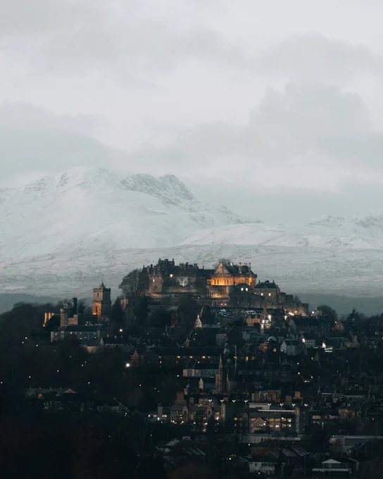 Castle on a hill seen above city buildings and below mountains covered in snow. 