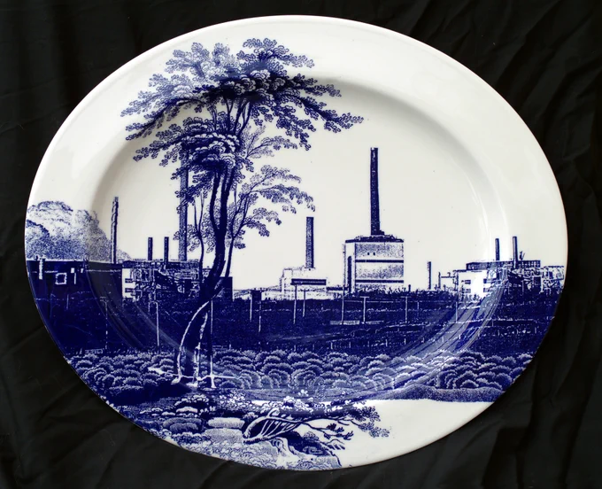 Oval ceramic plate featuring a blue and white illustration of a pastoral scene juxtaposed with industrial elements, including factories with smokestacks.