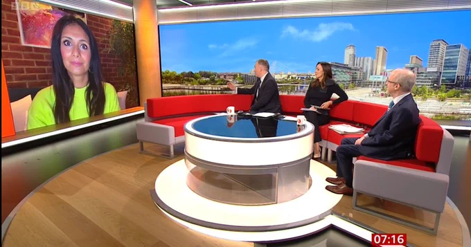 Inside a television studio, three people are seated around a curved desk with a city skyline backdrop, while Dr Anisha Patel is visible on a screen, participating remotely. The time display on the screen shows 07:16.