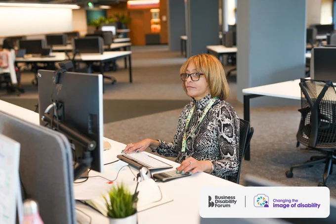 Person with less-visible disabilities uses an adaptive keyboard and calculator sitting at a desk in an office. The Business Disability Forum 'Changing the image of disability' logo is in the lower right corner of the image.