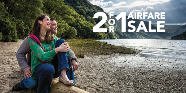Two people sit on a log on the beach together. Text overlaid reads "2 FOR 1 AIRFARE SALE."