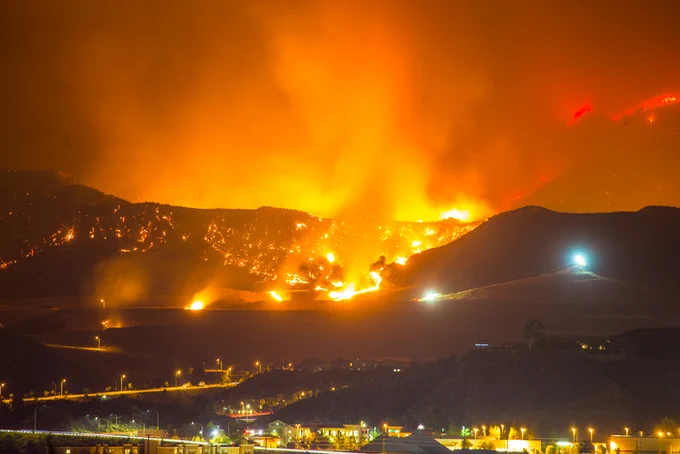 A nighttime image showing a large wildfire illuminating the sky in bright orange hues with visible flames engulfing a mountainside. In the foreground, city lights are visible under a smoky sky.