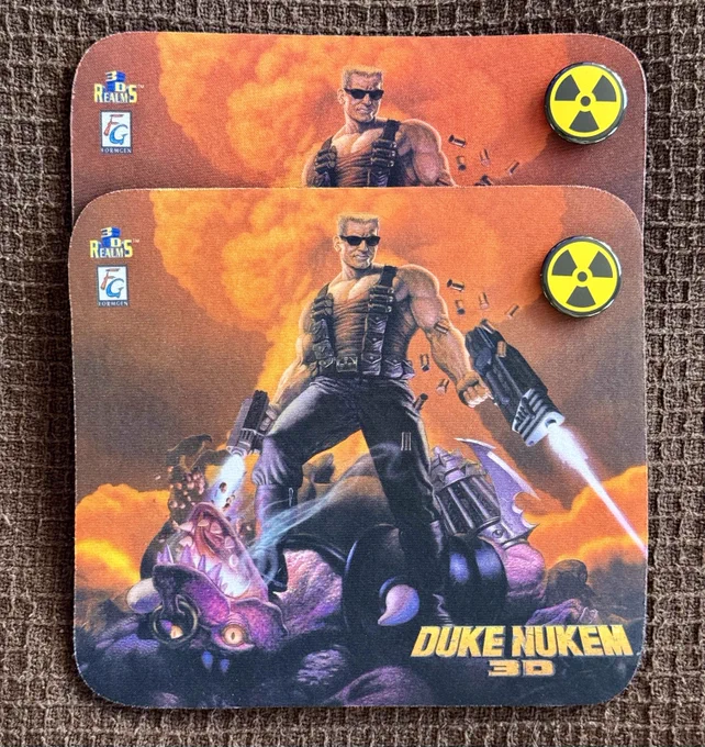 A photo of two vintage Duke Nukem 3D mouse pads featuring the iconic cover art of Duke standing triumphantly over an alien. Positioned on the top-right corners of the mouse pads are two circular Duke Nukem-themed magnets with the recognizable yellow radiation hazard symbol. The items are displayed on a textured brown surface, highlighting their retro charm and collector’s appeal.