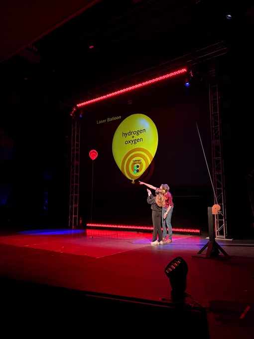 A presenter and a student aim a laser pointer at a balloon on a theater stage.