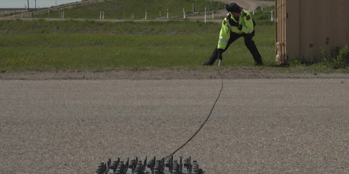 A police officer wearing a high-visibility jacket prepares to deploy at Tire Deflation Device. 