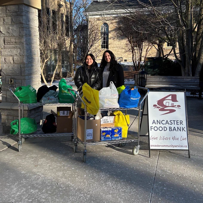 Photo of two women standing with carts of non-perishable food donations and the Ancaster Food Bank sandwich board.
