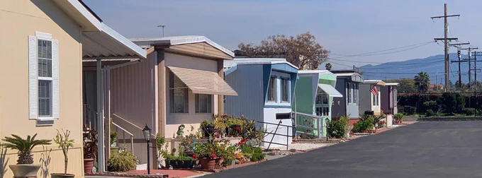 a row of pastel-colored mobile homes, in front of them, an open asphalt space. A cloudless blue sky, power wires stretching off into the distance.