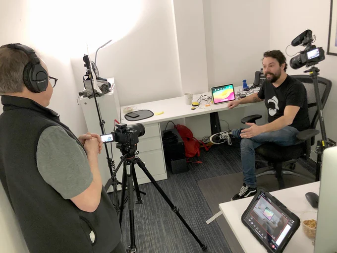 A person stands behind a video camera that is  directed to a man sitting at a desk.