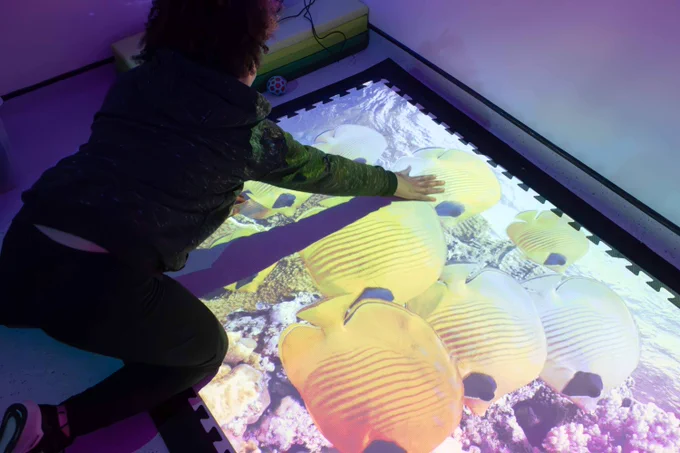 A child leans over an interactive pad on the floor. They put one hand on the pad, which shows fish swimming.