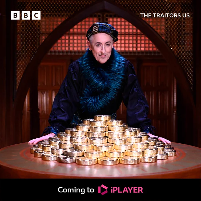 Alan Cumming stands over a desk of gold, wearing a purple robe.