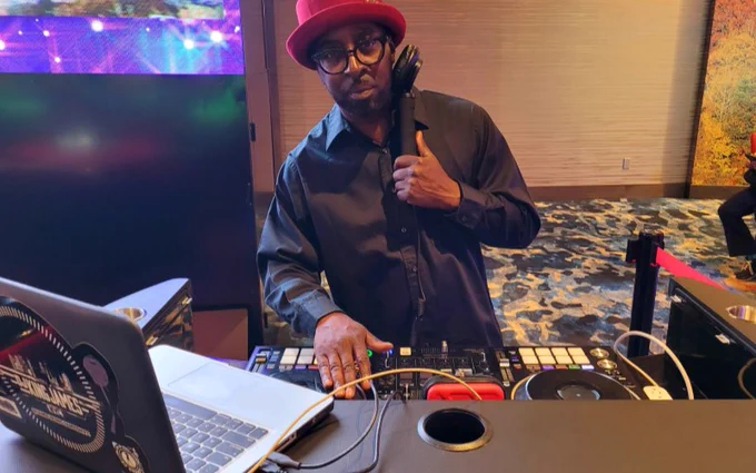 A person wearing a black shirt and a hat stands at a DJ booth, working on a mixer and holding headphones.
