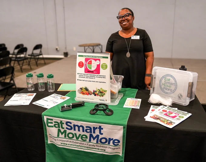 A Nutrition Education Facilitator stands by an educational table promoting the Eat Smart Move More program