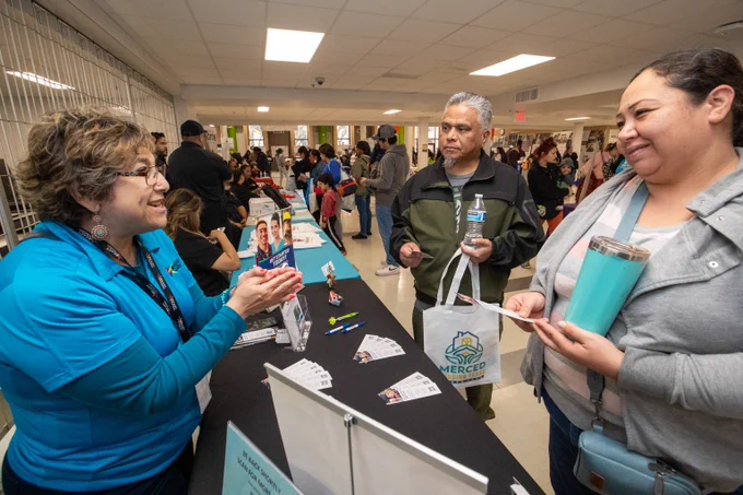 Two people speaking to each other at a table with various stationery at a public gathering.