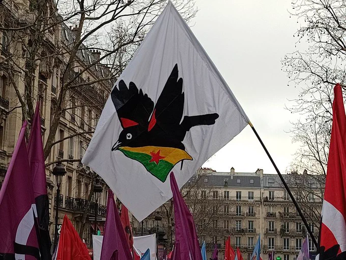 Alors que la manif s'élance, au centre un grand drapeau sur fond blanc avec le merle UCL tient le drapeau du Rojava dans son bec surmonte une demi douzaine de drapeaux UCL.