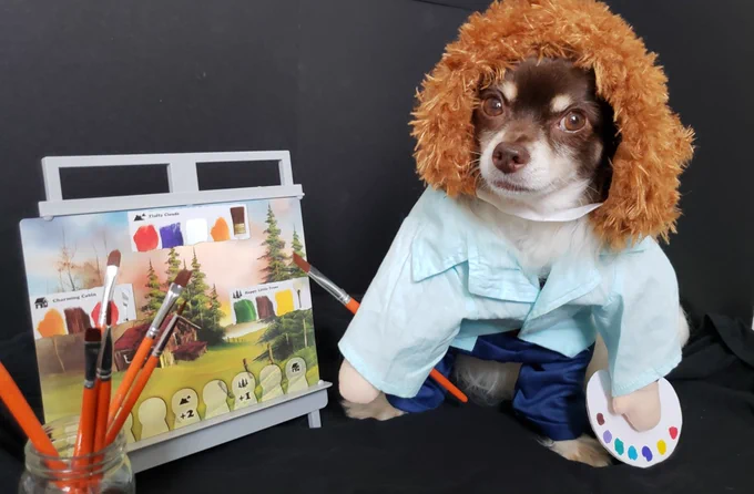 A dog in a Bob Ross costume sitting next to a painting.