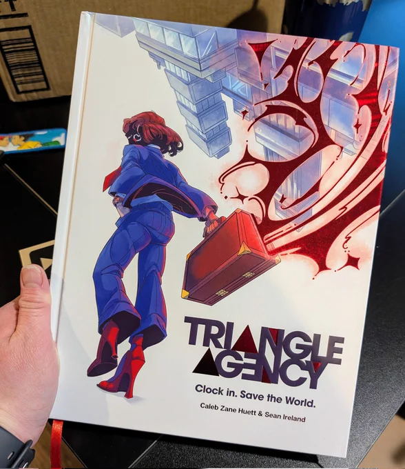 Triangle Agency book being held in one hand in front of a black table. 