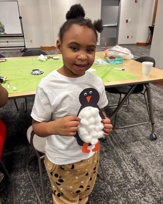 A child holding a craft project shaped like a penguin made with cotton balls, in a room with tables and crafting supplies.