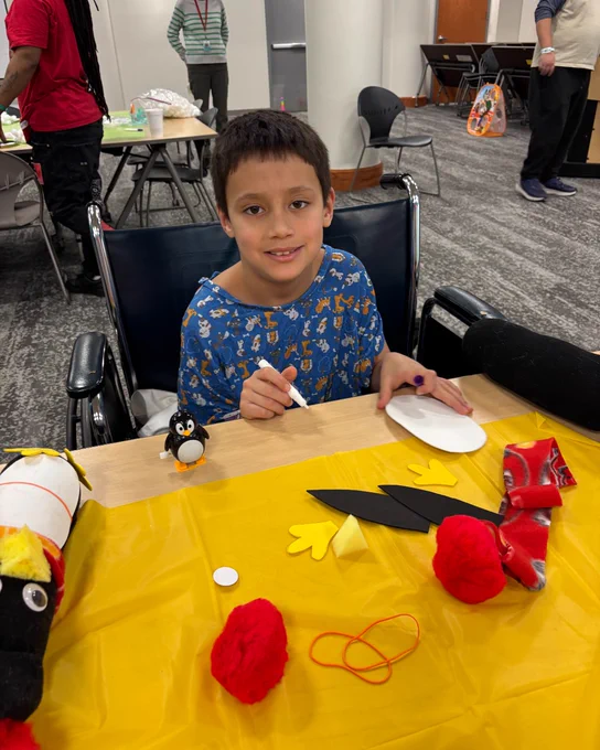 A child sits at a table covered with a yellow tablecloth, engaging in a crafts activity with various materials like yarn and paper laid out in front of them. There are handmade toys visible on the table.