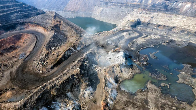 An aerial view of a large open-pit mining site with winding dirt roads, excavated rock layers, and machinery operating on the ground. Pools of water, scattered debris, and rising dust highlight the active mining operations.