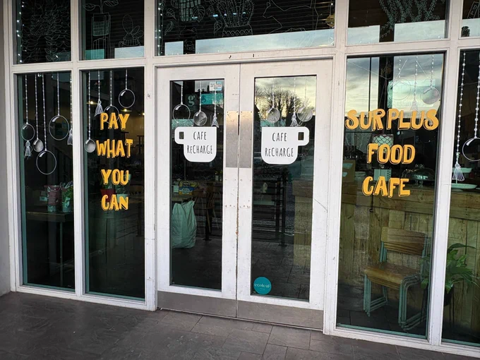Photo of a café. On the doors & windows the words "Pay as much as you can" and "surplus food café"