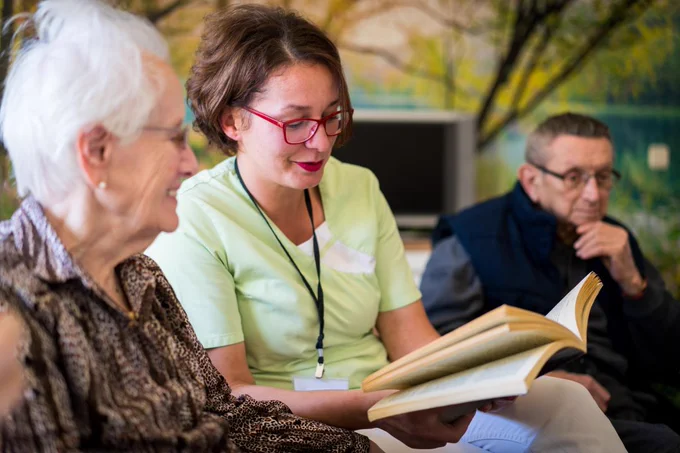 Photo of a woman reading a book with an older woman, who is smiling. There is an older man sitting next to them.