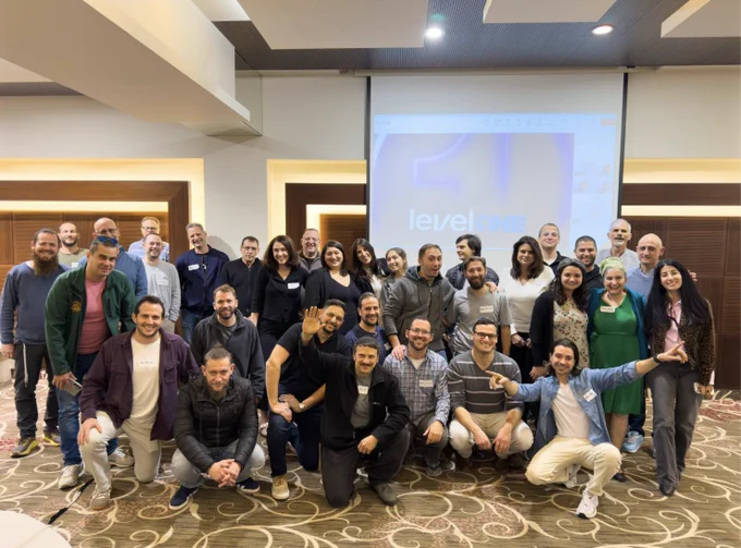 A group of around 30 UserWay and Level Access employees are gathered indoors, smiling and posing for a team photo. The background features a projector screen displaying the text "levelONE." The setting has modern decor, and the atmosphere feels positive and collaborative.