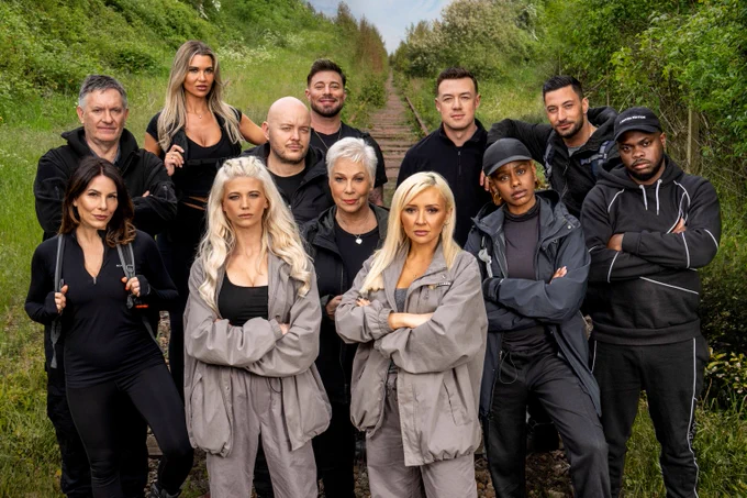 A group of twelve people dressed in black and grey tracksuits with rucksacks are stood looking to camera with a stealthy expression. They're standing outside in a field.