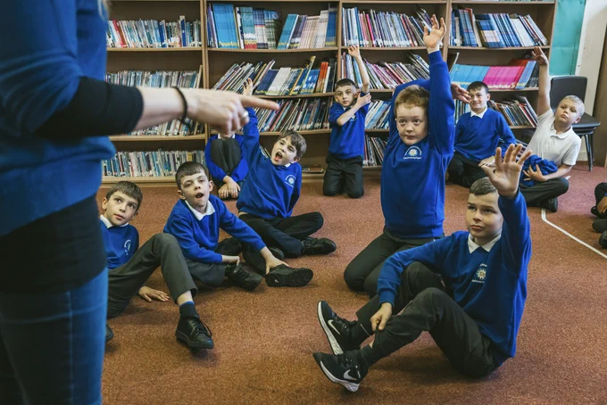 A teacher stands in front of a group of young students seated on the floor of a library. The students, dressed in blue sweaters, eagerly raise their hands to participate in the discussion. Behind them, shelves filled with books are visible.