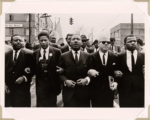 A black and white photograph mounted on an off-white mat. The photograph depicts Dr. Martin Luther King Jr.  locking arms with John Lewis, Reverend Jesse Douglas, James Forman and Ralph Abernathy as they march down a street in Selma, Alabama.
