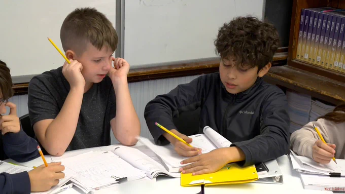A group of middle school students sit together at a table working on math problems from a workbook.