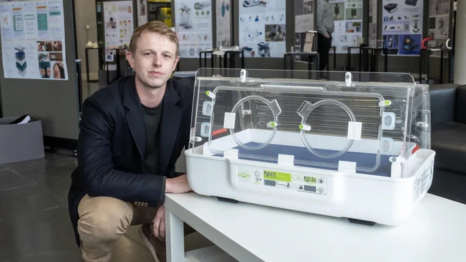 James Roberts MBE sits next to the mOm Incubator placed on a white table. 