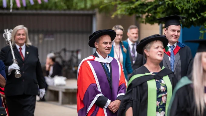 Sir Warren East FRS FREng is dressed in Honorary Degree robes and is walking as part of the procession at graduations. Around him are others in the procession.