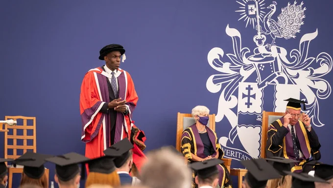 Professor Ivan Browne OBE stands on the Loughborough University graduation stage. He is dressed in honorary graduate robes and behind him is a purple backdrop with the Loughborough logo and two individuals sat down. 