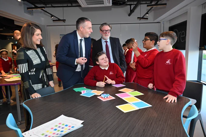 Three adults talking to three pupils in red jumpers who have coloured squares and small blue Indi robot on the table on front of them. 
