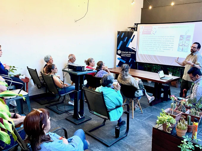 A man stands and speaks to a seated class with slides on a screen behind him. 