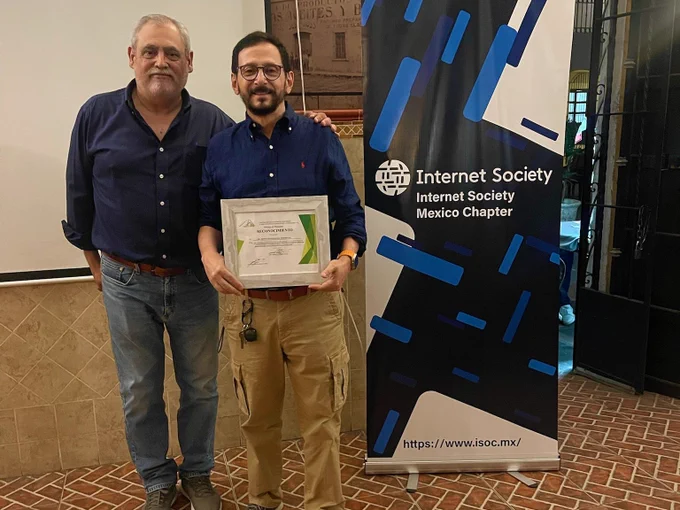 Two men stand together in front of an Internet Society Mexico Chapter sign. 