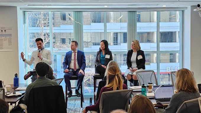 Two men and two women sit on stools at the front of a room presenting to a crowd during a professional conference.