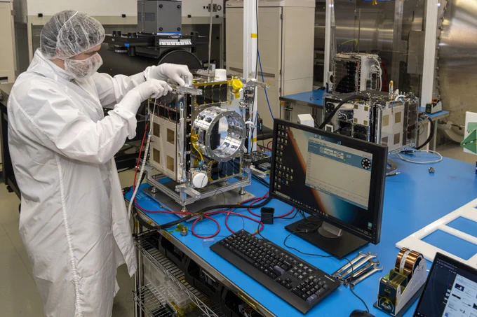 A person wearing a white laboratory coat, gloves, and a hair net uses a hand-held tool to adjust a satellite on a table, while looking at a computer screen, in a laboratory.