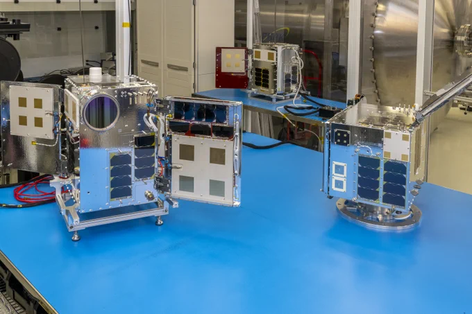 Three metal box-shaped satellites on a table in a laboratory.