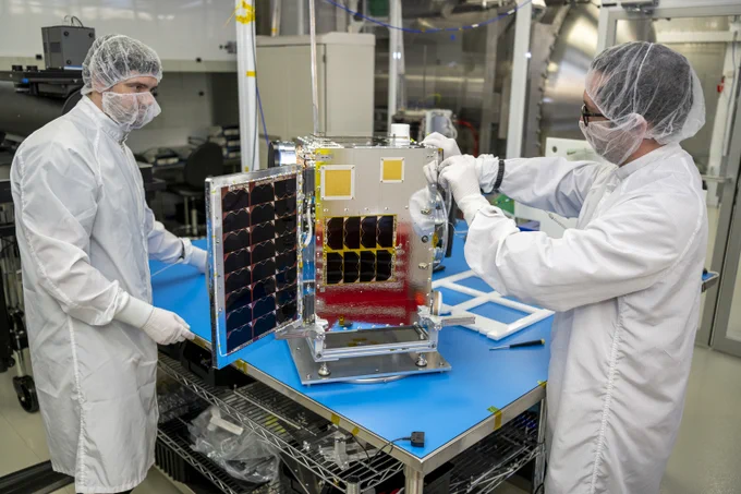 Two people wearing white laboratory coats, gloves, and hair nets work on a satellite on a table between them in a laboratory.