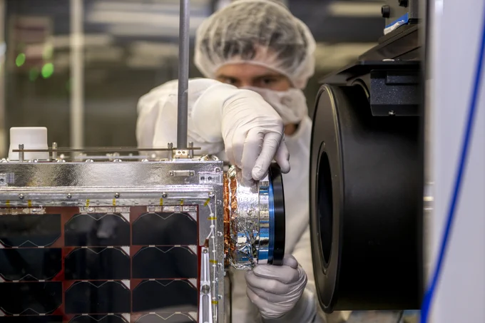 A person wearing a white laboratory coat, gloves, and a hair net adjusts a cylindrical object on the side of a small satellite inside a laboratory.