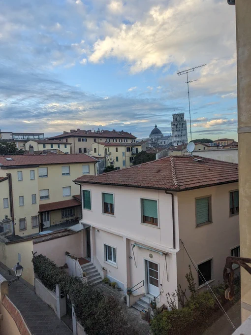 View of an Italian, Tuscan cityscape from a window; in the distance can be seen the Leaning Tower of Pisa and the dome of the cathedral, below a late afternoon blue sky with clouds.