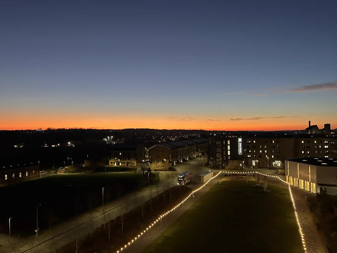 Image shows a sunset view of Northampton from the westerly facing balcony of the Senate Building at the University of Northampton. In the foreground is an area of grass with spring lights around it at a height of 12 feet. To the right of the grass area is the Learning Hub of the University. To the left of the grass area is a road on which there is a bus at a bus stop. In the centre right of the photograph is the Sunley Hotel with the name illuminated on the right hand side of the building. The remainder of the photograph shows Northampton at sunset with various building and street lights illuminated.