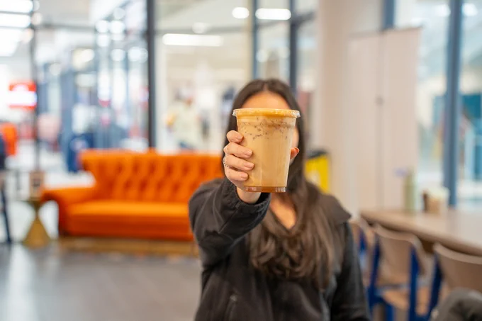 Person holding iced coffee in focus with a blurred background of an indoor cafe setting.