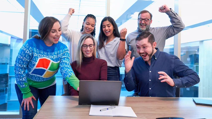 A group of six people gathered around a laptop, celebrating with joyful expressions. One person is wearing a colorful sweater with the Windows logo, while others are smiling, cheering, and raising their hands in excitement. 