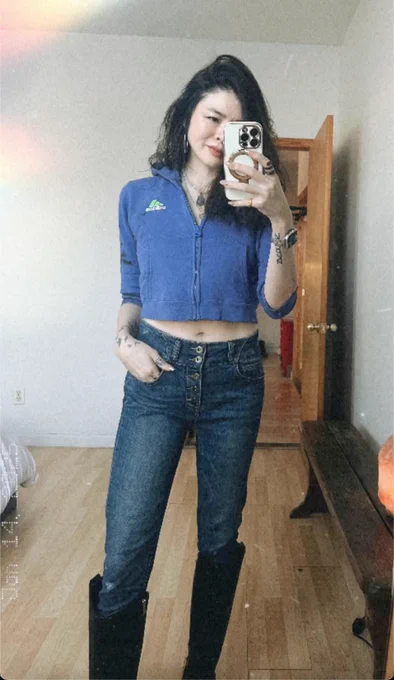Ruibo QIan stands in front of a mirror taking a selfie in black boots, jeans and a blue croptop.